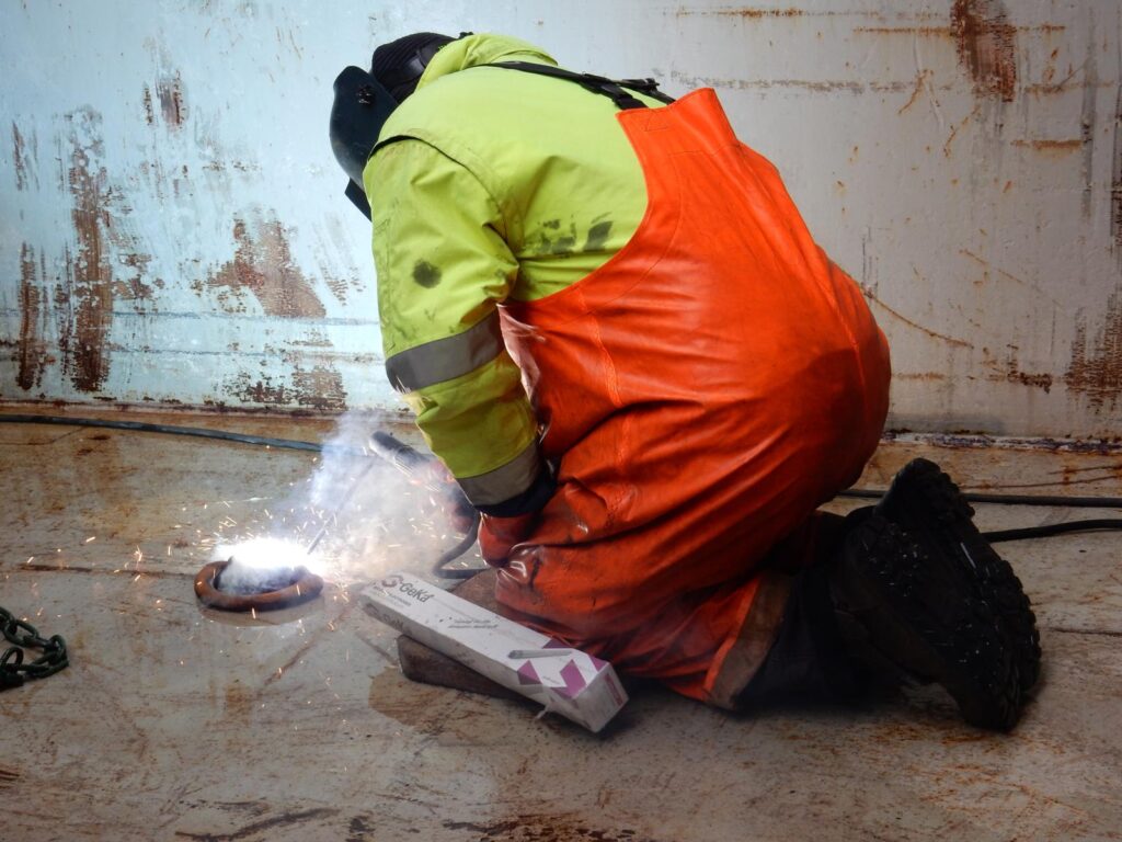 Foto eines Arbeiters in gelber und orange Sicherheitskleidung beim Schweißen mit Schweißmaske und sichtbaren Funken für Schwerlast-Metallarbeiten
