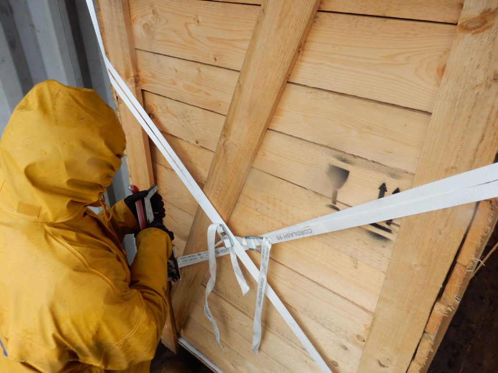 Arbeiter in gelber Schutzkleidung sichert Holzkiste mit Spanngurten – transporttechniker.de 