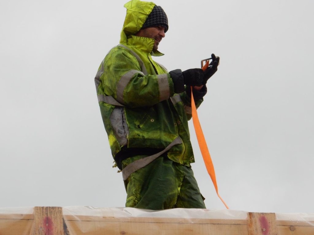 Foto eines Arbeiters in grüner Regenkleidung mit Kapuze und grauer Mütze bei Sicherungsarbeiten: Er hält einen orangefarbenen Gurt und bedient ein Gerät auf einer Holzkiste