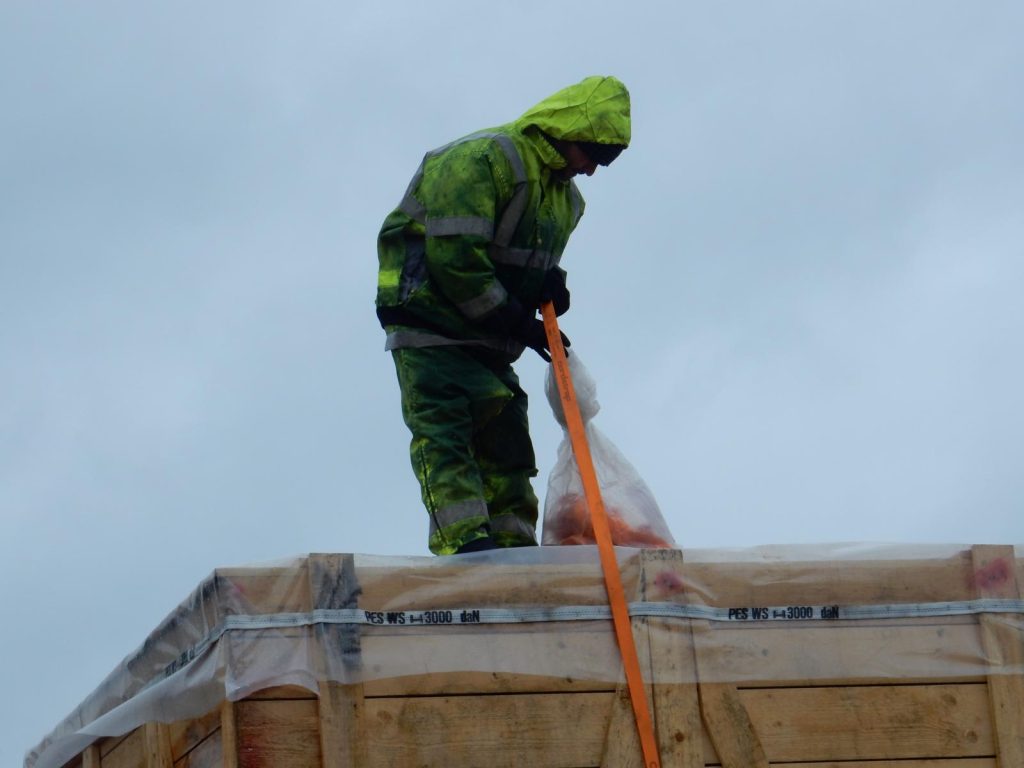 Foto eines Arbeiters in grüner Regenkleidung mit Kapuze bei Sicherungsarbeiten: Er steht auf einer mit Plane abgedeckten Holzkiste (Beschriftung "PES WS 3000 daN") und hält einen orangefarbenen Gurt