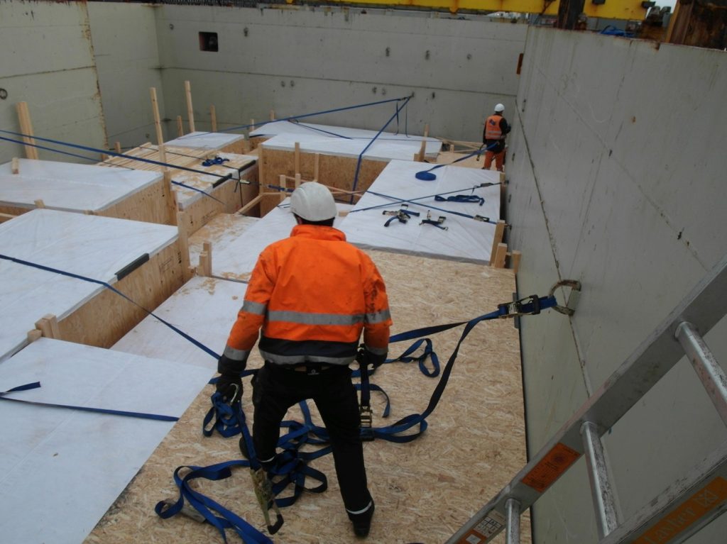 Foto eines Arbeiters in orange Sicherheitskleidung und weißem Helm mit blauem Sicherheitsgeschirr bei Montagearbeiten auf einer Plattform aus OSB-Platten und Metall für Schwerlastkonstruktion