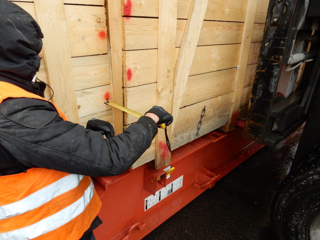 Foto eines Arbeiters in orange Warnweste und dunkler Kleidung mit Kapuze bei Kontrollmessungen mit gelbem Messband an einer Holzkiste mit roten Markierungen für Schwerlast-Qualitätskontrolle