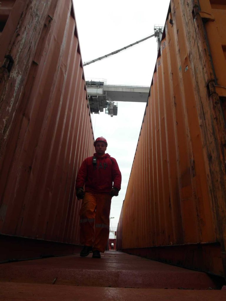 Foto eines Arbeiters in roter Sicherheitskleidung und rotem Helm beim Gehen durch eine Gasse zwischen gestapelten Containern mit Container-Kran im Hintergrund für Schwerlast-Logistik
