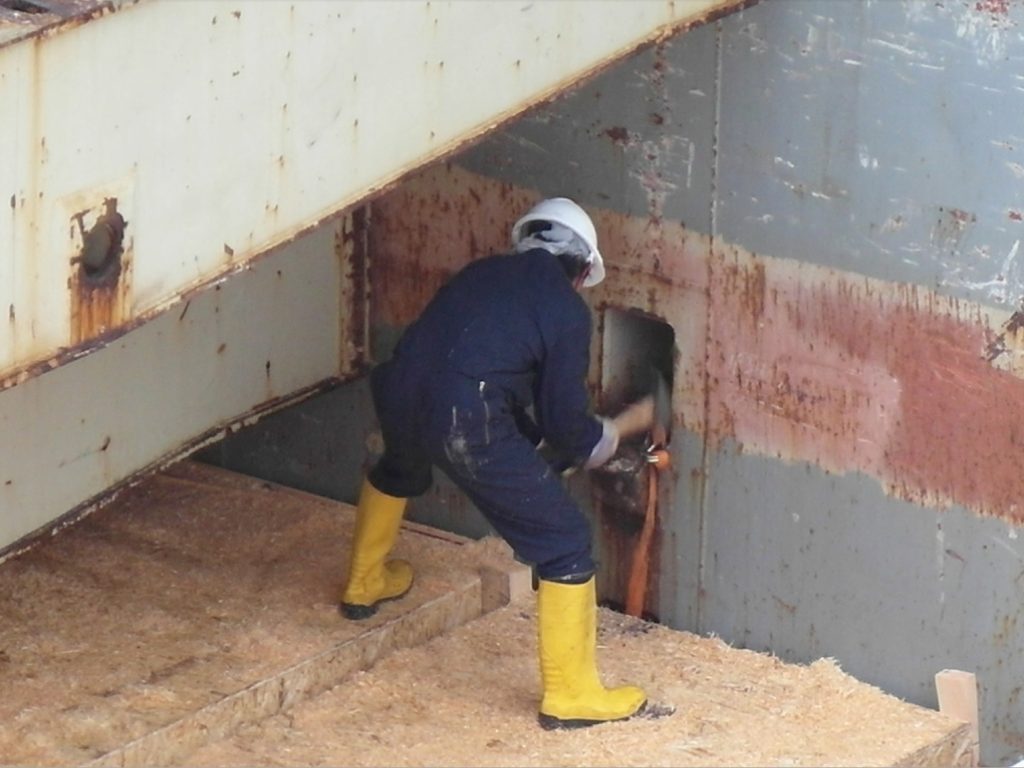 Foto eines Arbeiters in dunkler Arbeitskleidung und weißem Helm mit gelben Gummistiefeln bei Schleifarbeiten an einer verrosteten Metallwand mit sichtbaren Funken für Schwerlast-Metallarbeiten