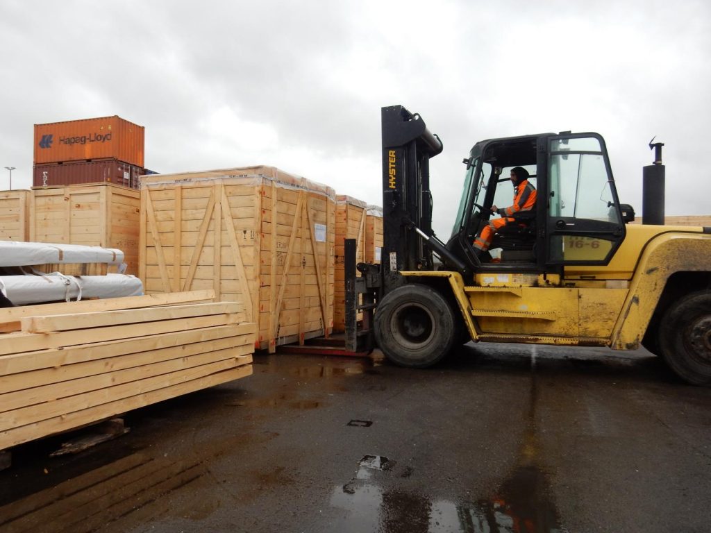 Foto eines gelben Hyster-Gabelstaplers, gesteuert von einem Arbeiter in orange Sicherheitskleidung, beim Transport von großen Holzkisten für Schwerlastgut mit gestapelten Hapag-Lloyd-Containern im Hintergrund