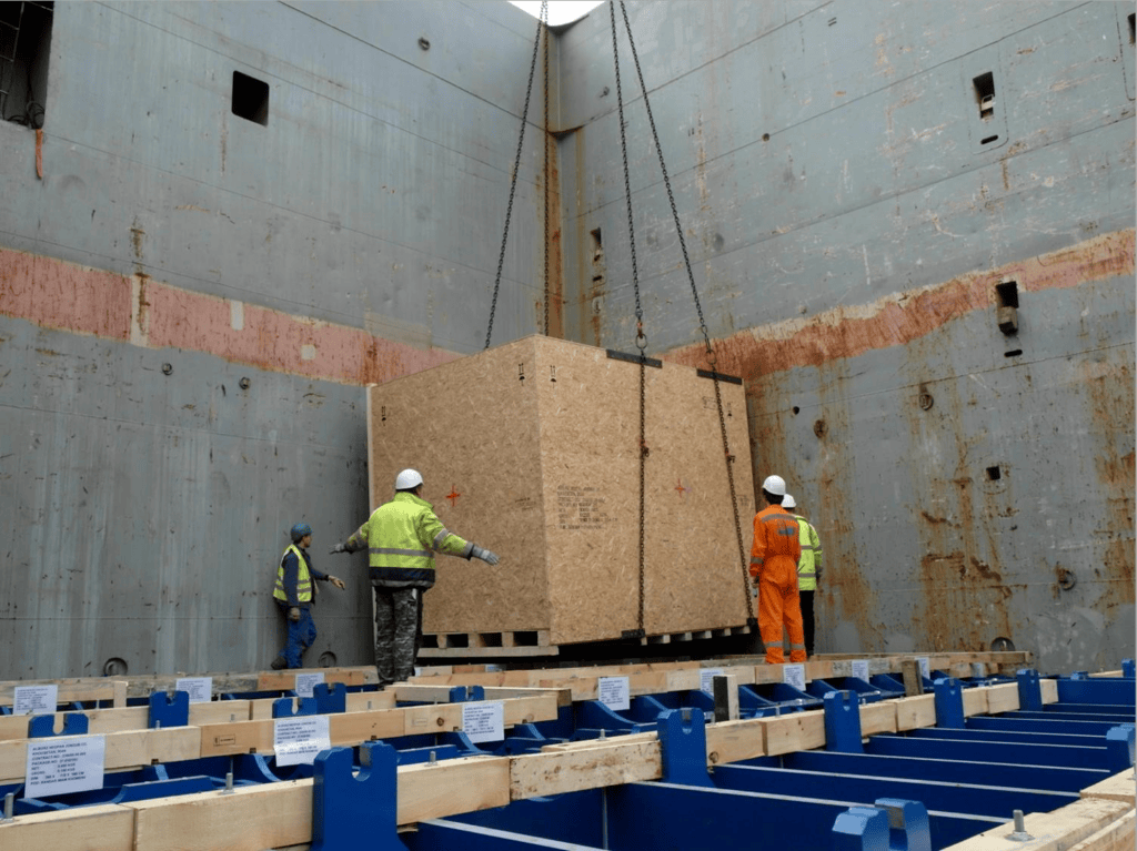 Foto einer gesicherten Holzkiste mit Schwerlastgut in Open-Top-Container (HDMU 321070 1): Kreuzweise mit Gurten verzurrt, Arbeiter in orange Sicherheitskleidung