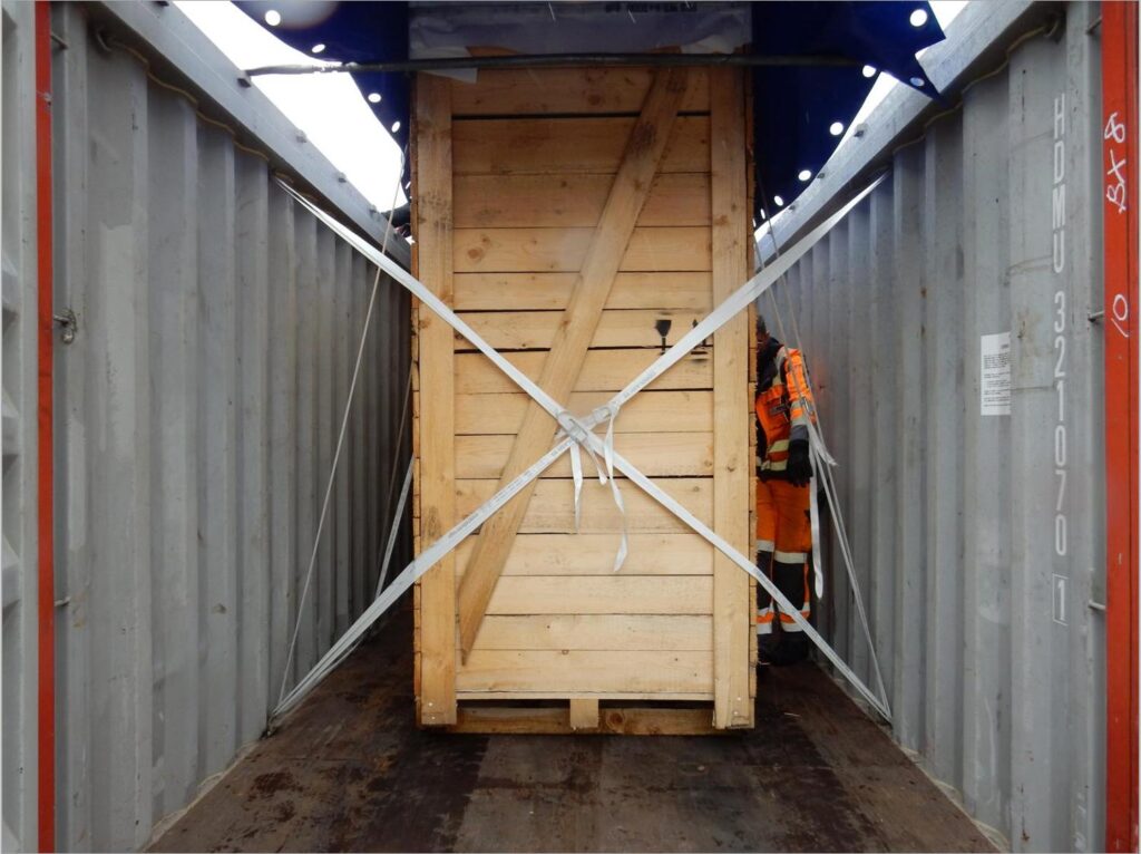 Foto einer gesicherten Holzkiste mit Schwerlastgut in Open-Top-Container (HDMU 321070 1): Kreuzweise mit Gurten verzurrt, Arbeiter in orange Sicherheitskleidung