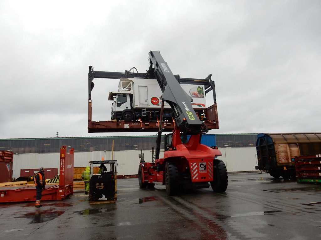 Foto eines roten KALMAR-Reachstackers beim Heben eines Containers mit LKW und "ECO efficient"-Aufschrift, zwei Arbeiter in orange Sicherheitskleidung im Hintergrund
