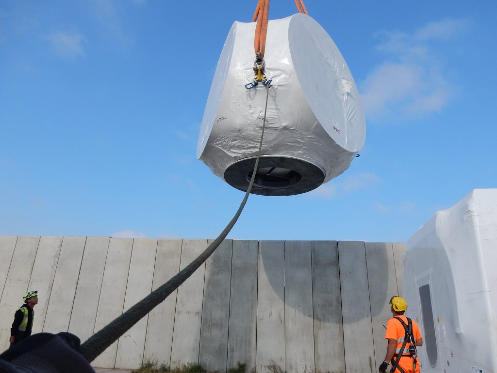 Foto eines in weiße Folie eingepackten zylindrischen Schwerlastobjekts, das mit orangefarbenen Gurten von einem Kran gehoben wird: Zwei Arbeiter in Sicherheitskleidung (gelber und grüner Helm) beaufsichtigen den Transport vor grauer Betonmauer