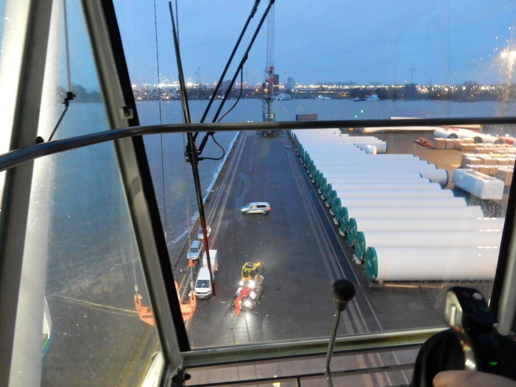 Blick aus dem Führerhaus eines Krans auf eine Werft – Transportfahrzeuge mit weißen Rotorblättern für Windkraftanlagen, Hafenbecken und Industrieanlagen im Hintergrund, klare Sicht auf die Arbeitsfläche