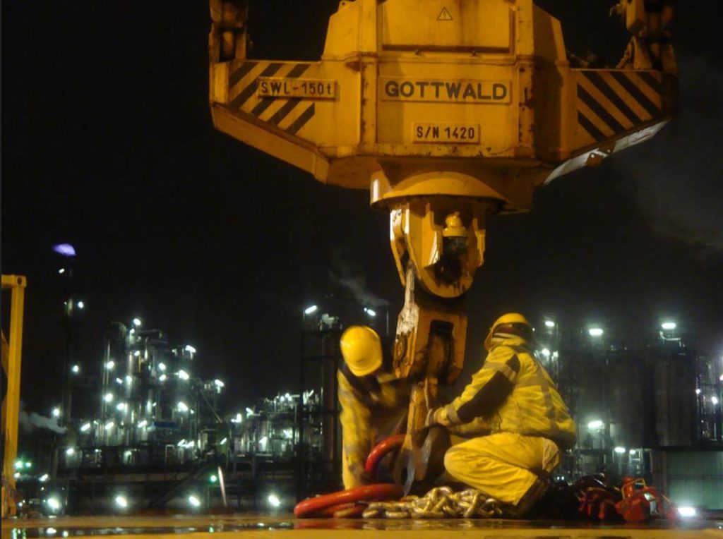 Fachkräfte beim Anschlagen einer Schwerlastladung unter einem Hafenkran während einer Nachtverladung in einer Industrieanlage im Bereich Schwergutumschlag und Projektlogistik