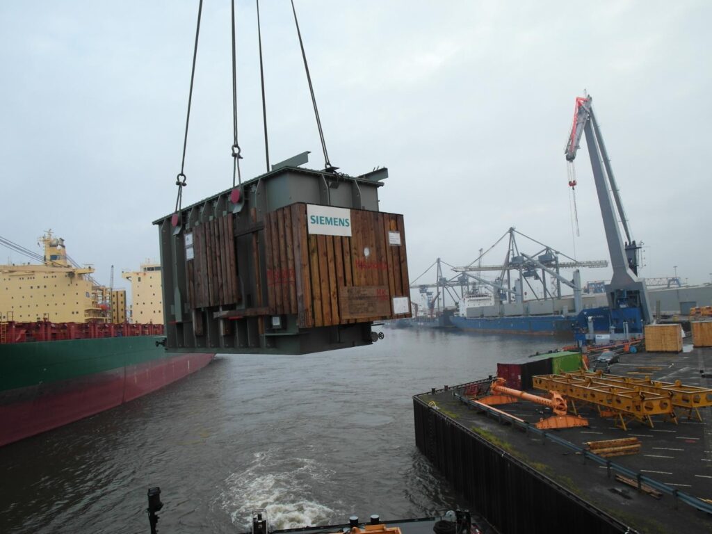 Schwerlasttransport eines Siemens-Transformators per Kran auf einem Schiff im Hafen – großer, verpackter Transformator wird von Kränen gehoben, im Hintergrund weitere Schiffe, Container und Hafeninfrastruktur