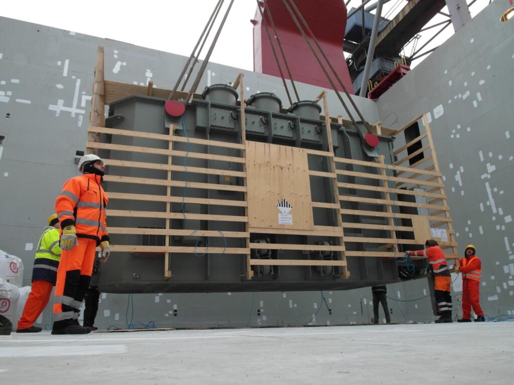 Foto vom Schwerlastumschlag: Transformator in Holzrahmen wird mit Kran von Arbeitern in orange Sicherheitskleidung verladen