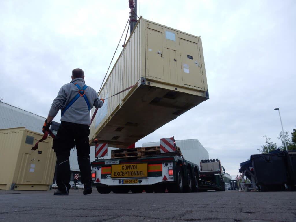 Foto eines Arbeiters in grauer Arbeitskleidung mit blauem Sicherheitsgeschirr beim Schwerlasttransport: Beiger Container wird mit Kran über einem LKW mit Aufschrift "CONVOI EXCEPTIONNEL" und "VOERTUIG ZWAAR UIT" schwebend bewegt
