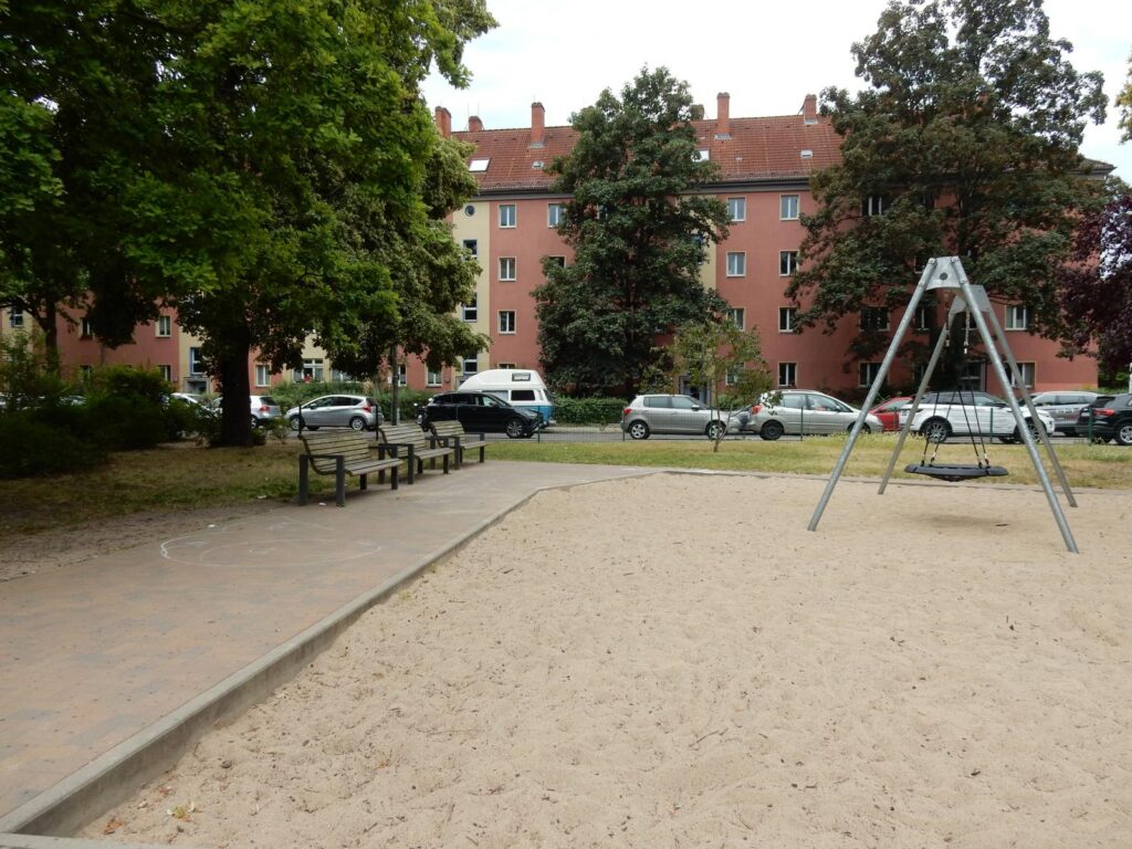 Spielplatz in der Dusekestraße, Berlin – Sandfläche mit Schaukel, Bänken und Wohnhäusern im Hintergrund – transporttechniker.de