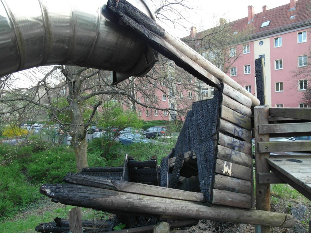 Spielplatz in der Dusekestraße, Berlin – Verbrannte Holzkonstruktion mit Rutsche und Wohnhäusern im Hintergrund – transporttechniker.de