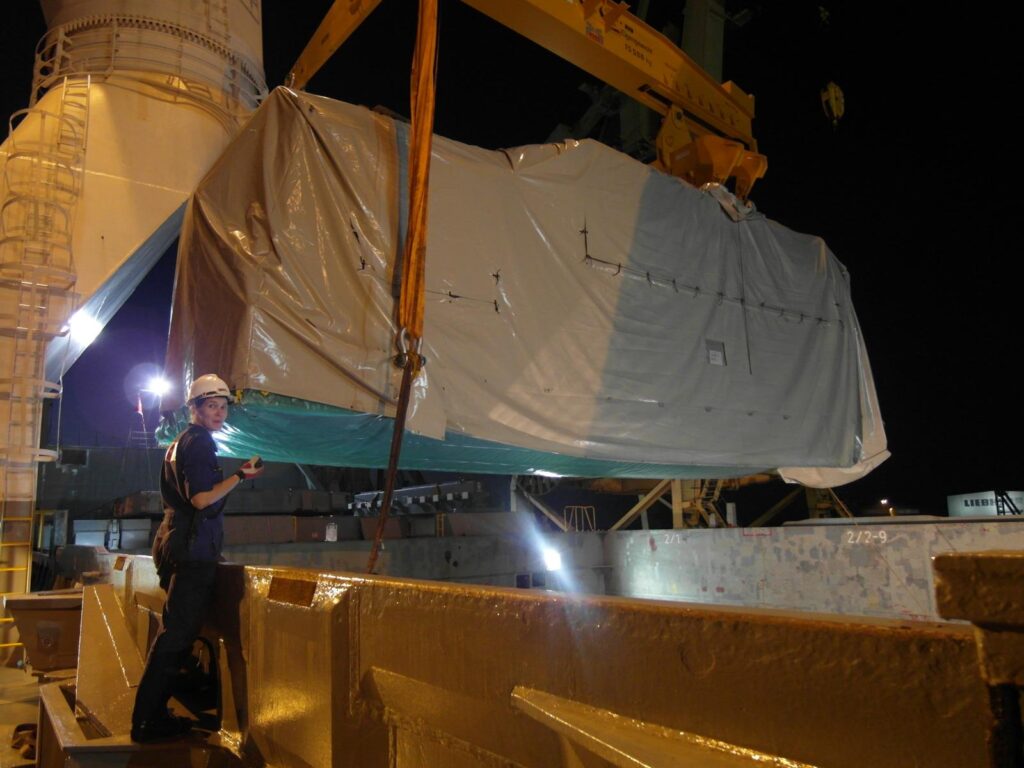 Foto einer Arbeiterin in blauer Arbeitskleidung und weißem Helm mit Gerät bei Nacht: Ein großes, in Plane eingepacktes Schwerlastobjekt wird von einem Kran gehoben, im Hintergrund ein weißes Schiff und weitere Krananlagen