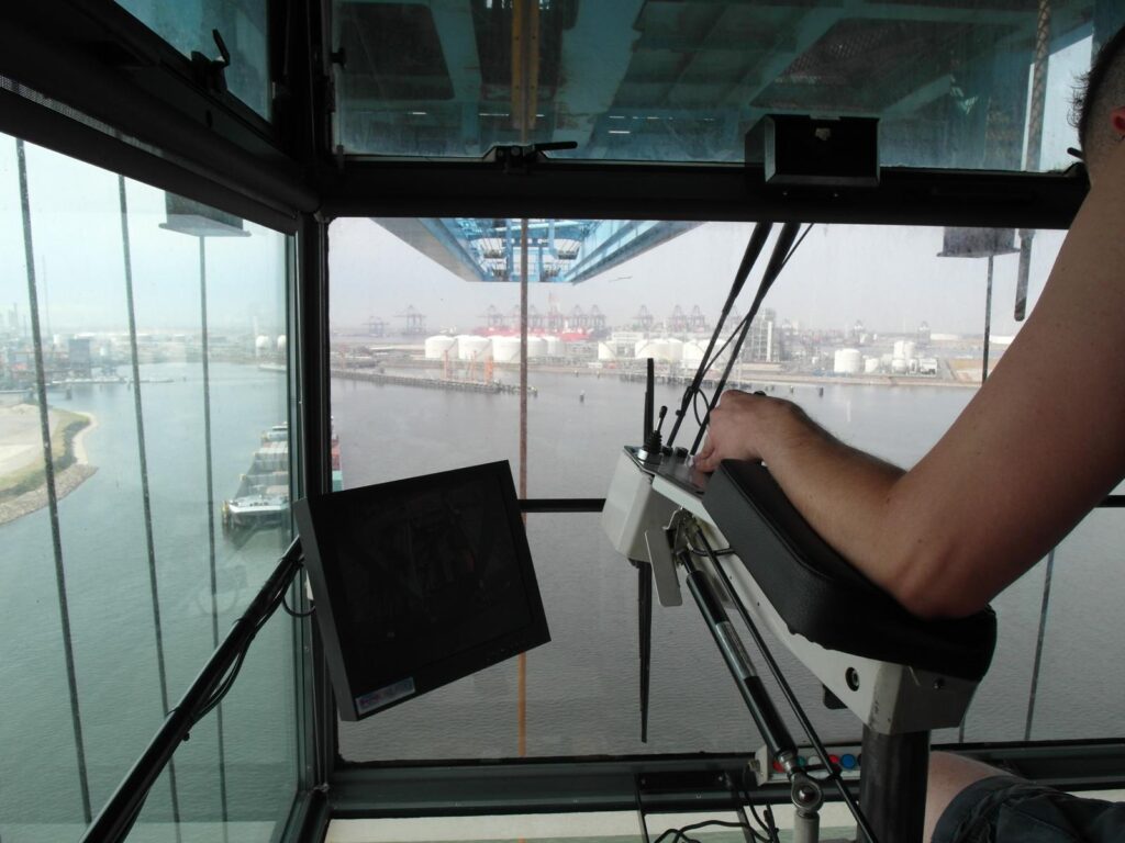 Foto aus der Kranführer-Kabine: Blick durch die Glasfront auf Hafen mit Kränen, Tanks und Wasser während des Schwerlastumschlags