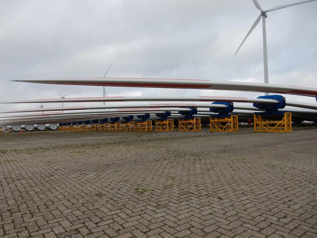 Gestapelte Windkraft-Rotorblätter auf gelben Lagergestellen mit Windrädern im Hintergrund – transporttechniker.de