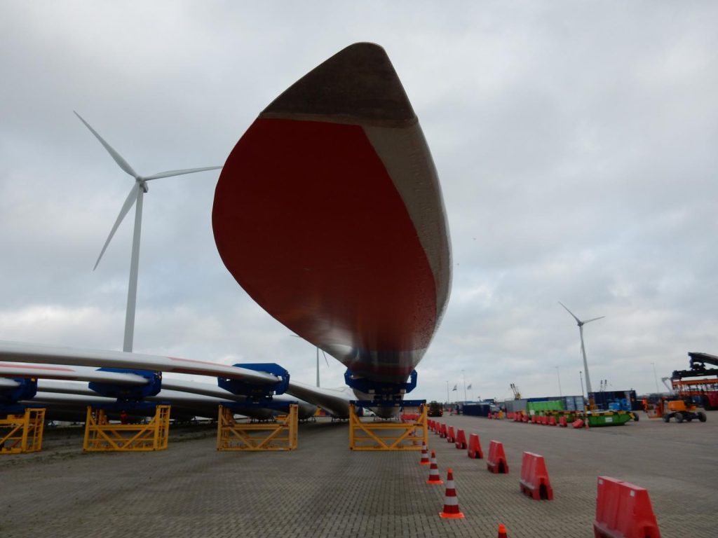 Rotes Rotorblatt einer Windkraftanlage in Hafenlager mit Windrädern im Hintergrund – transporttechniker.de